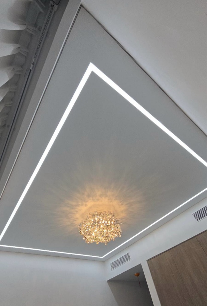 Minimalist bespoke stretch ceiling featuring integrated linear LED lighting surrounding a decorative chandelier in a luxury villa. Minimalist bespoke stretch ceiling featuring integrated linear LED lighting surrounding a decorative chandelier in a luxury villa.