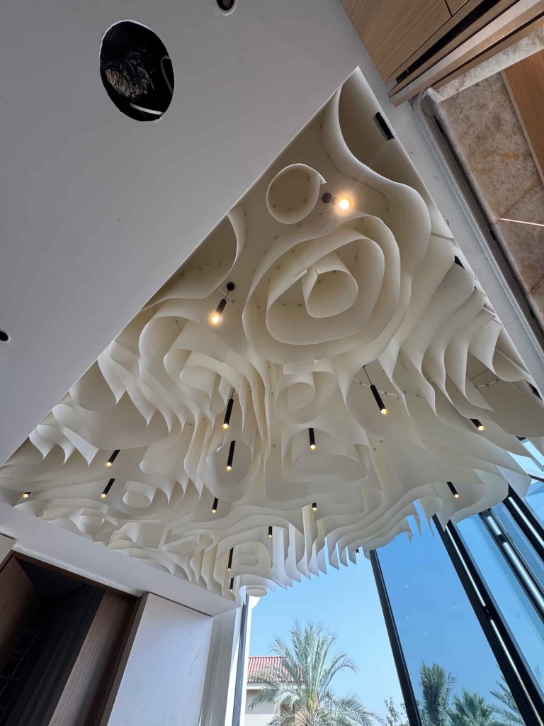 Wide-angle view of a luxury villa featuring a bespoke fabric ceiling with organic flowing shapes and integrated pendant lighting. Designed exclusively for this residence, the ceiling serves as the main architectural focal point while enhancing the spacious contemporary interior. Wide-angle view of a luxury villa featuring a bespoke fabric ceiling with organic flowing shapes and integrated pendant lighting. Designed exclusively for this residence, the ceiling serves as the main architectural focal point while enhancing the spacious contemporary interior.