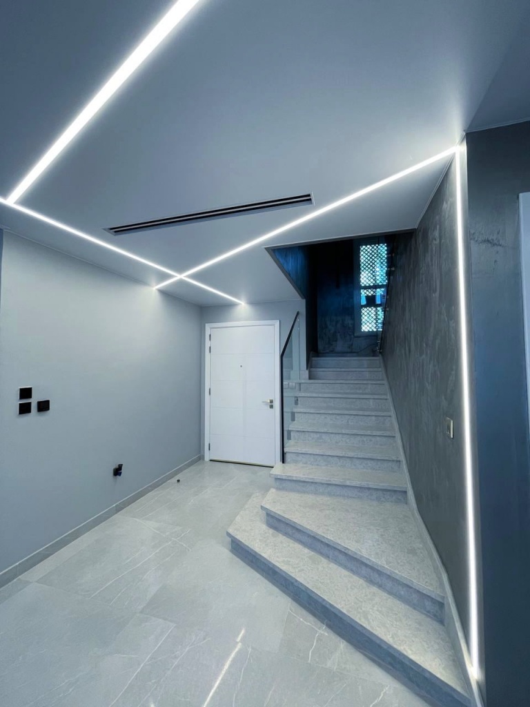 Minimalist stretch ceiling featuring continuous linear lighting that extends across the ceiling and walls. Minimalist stretch ceiling featuring continuous linear lighting that extends across the ceiling and walls.
