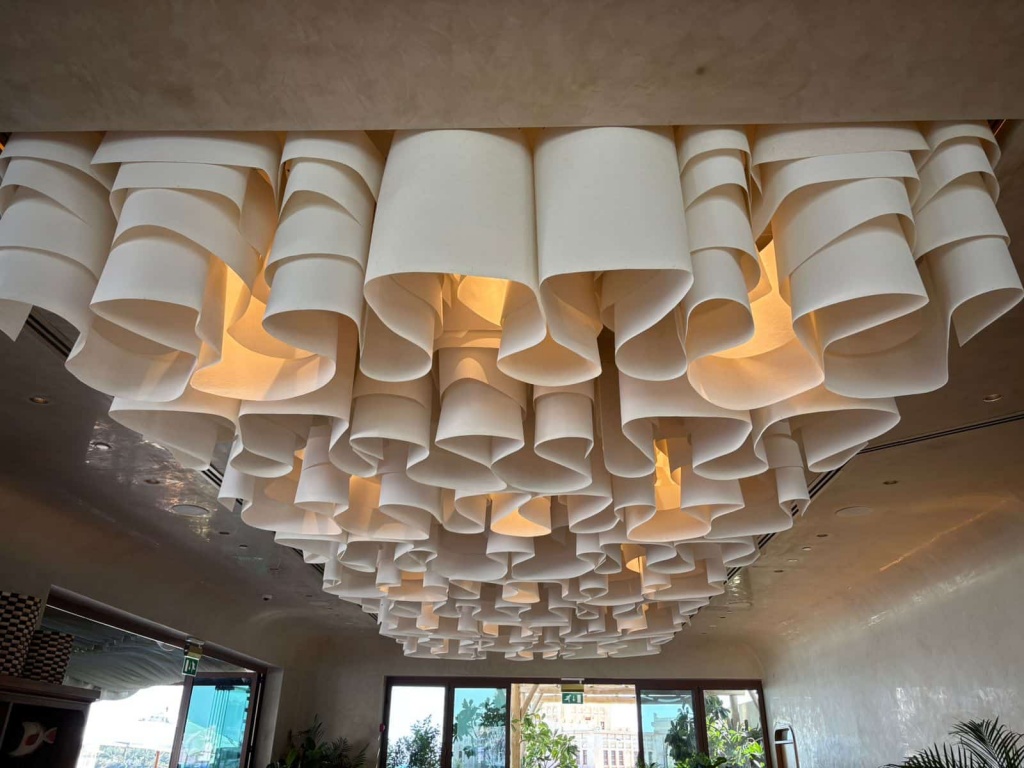 Large rectangular sculptural ceiling formed from layered white fabric waves and integrated lighting in a premium architectural interior. Large rectangular sculptural ceiling formed from layered white fabric waves and integrated lighting in a premium architectural interior.