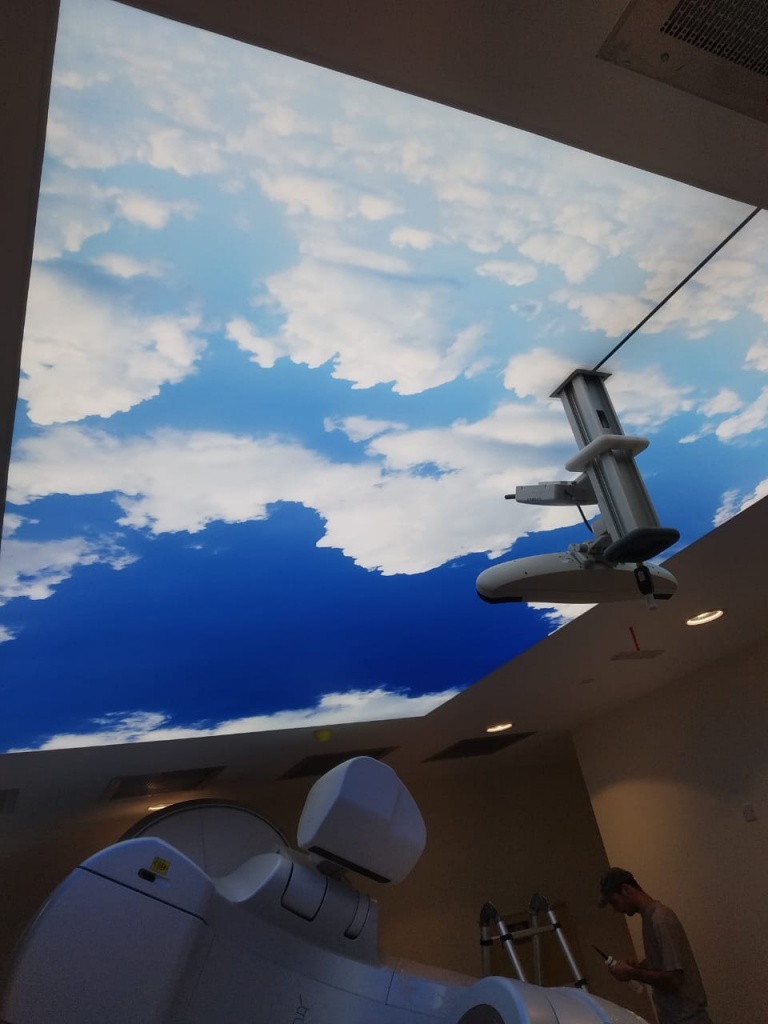 Custom printed stretch ceiling with blue sky and clouds integrated with ceiling-mounted medical equipment in an Abu Dhabi healthcare facility. Custom printed stretch ceiling with blue sky and clouds integrated with ceiling-mounted medical equipment in an Abu Dhabi healthcare facility.