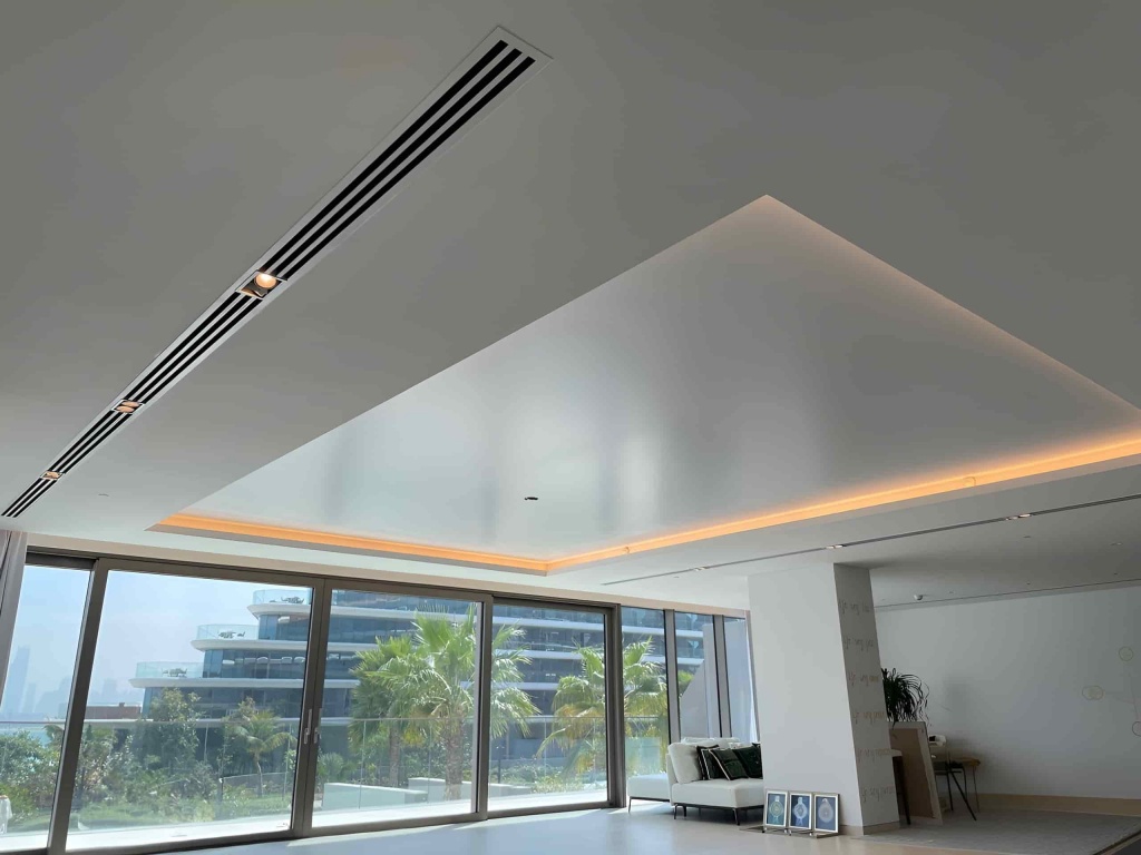 Large white stretch ceiling feature with warm perimeter LED illumination and integrated linear AC grilles in a modern Dubai villa. Large white stretch ceiling feature with warm perimeter LED illumination and integrated linear AC grilles in a modern Dubai villa.